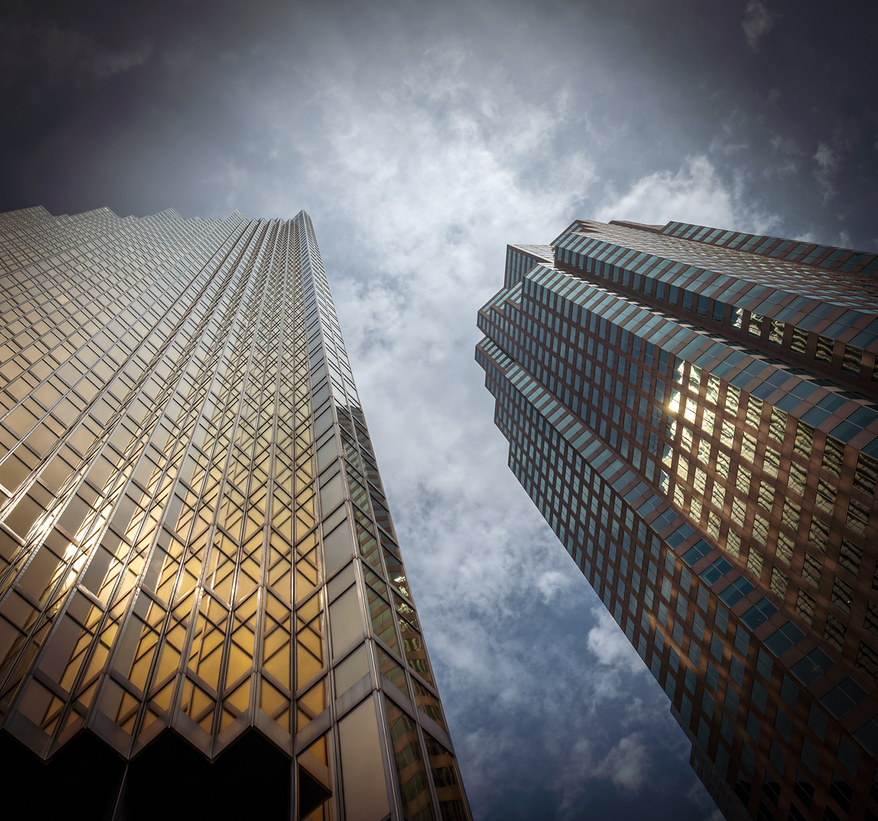 Two glass skyscrapers loom upward toward a cloudy sky, shot from ground level.